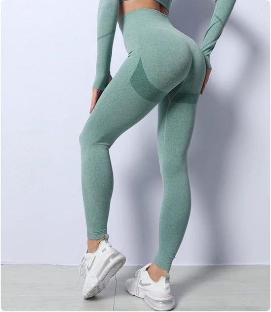 Vrouw in groene sportkleding, waaronder Zie er geweldig uit en voel je geweldig in deze 3-delige yoga workoutset, en witte sneakers poserend tegen een grijze achtergrond, waarbij de nadruk ligt op haar fitnesskleding.