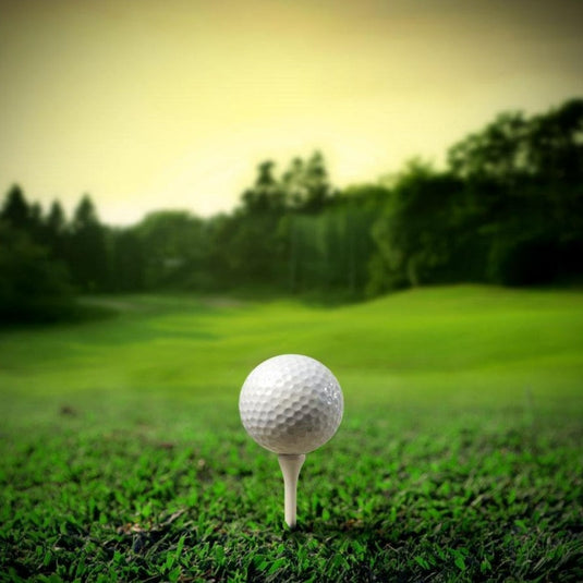 Golfbal op een professionele houten golftee in een grasveld met een onscherpe achtergrond van bomen en een warme zonsonderganghemel.