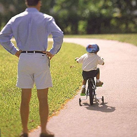 Een man ziet een jong kind met een helm op een kleine fiets met Zijwieltjes voor kinderfietsen over een zonnig parkpad rijden, waardoor de veiligheid gegarandeerd is.