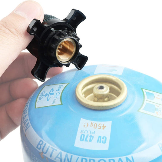 Een persoon houdt een zwarte Campingaz-adapter vast naast een blauwe butaangasfles, ter voorbereiding op het aansluiten van de adapter voor het koken tijdens zijn kampeeravontuur.