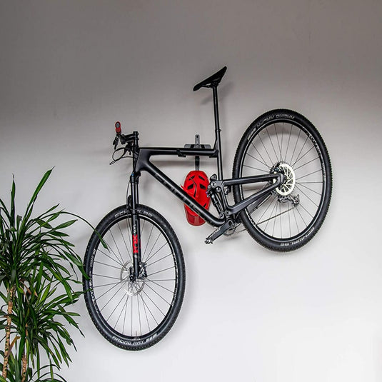 Zwarte mountainbike gemonteerd op een grijze muur met behulp van Optimaliseer je ruimte met onze stijlvolle fiets ophangsysteem, met een rode helm aan het stuur en een groene plant aan de linkerkant.