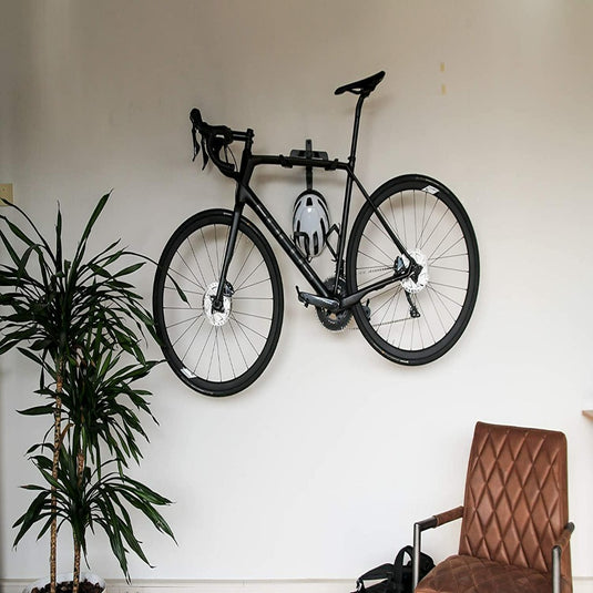 Een zwarte racefiets gemonteerd op een witte muur boven een bruinleren stoel met links een grote potplant met behulp van een ruimtebesparend Optimaliseer je ruimte met onze stijlvolle fiets ophangsysteem.