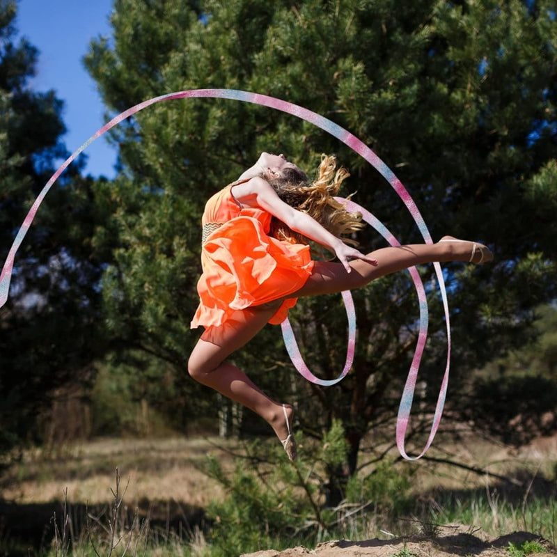 Laad afbeelding in Galerijviewer, Een jonge vrouwelijke turnster die een sprong maakt met een lint in een zonnige buitenomgeving, gekleed in een oranje jurk die geschikt is voor ritmische sportgymnastiek, en gebruik maakt van Dansbanden voor kinderen om haar routine te verbeteren.
