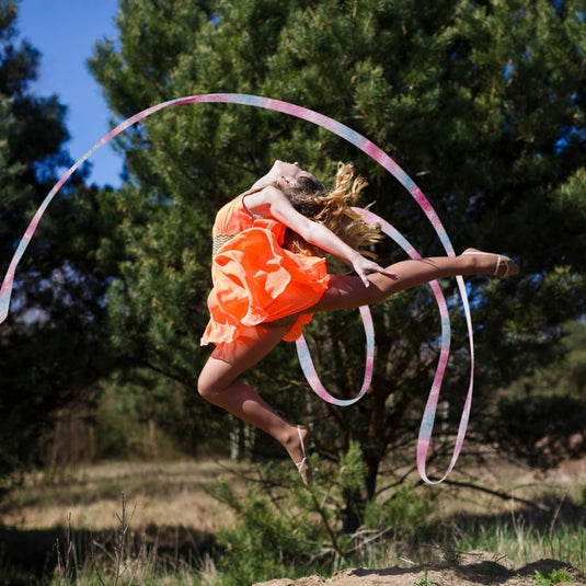Een jonge vrouwelijke turnster die een sprong maakt met een lint in een zonnige buitenomgeving, gekleed in een oranje jurk die geschikt is voor ritmische sportgymnastiek, en gebruik maakt van Dansbanden voor kinderen om haar routine te verbeteren.