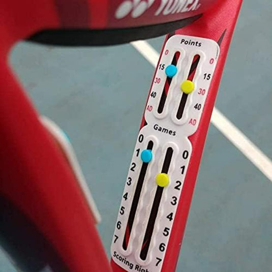 Close-up van een Draagbaar tennis scorebord op een rood racket, met punten en spellen met gekleurde pinnen op genummerde tracks. De baanlijnen zijn zichtbaar op de achtergrond, wat de compatibiliteit met elk universeel montagesysteem laat zien.