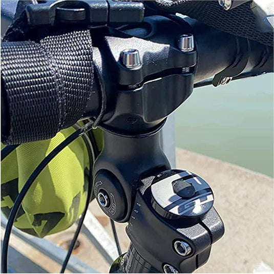 Close-up van een fietsstuur en stuurpen, met de schakelhendel en bevestigingsklem, met een glimp van een groene tas en water zichtbaar op de achtergrond. De Verstelbare stuurpen benadrukt zijn aanpassingsvermogen voor comfortabele fietsen.