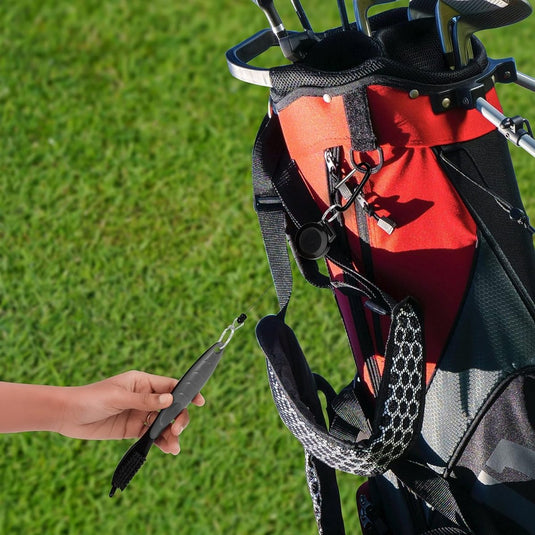 De hand van een golfer reikt naar een golfclub uit een tas op een groen gazon, naast een Golfclubborstel en groove cleaner: houd je clubs in topconditie.