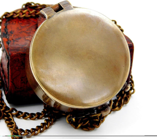 Antiek koperen medaillon aan een ketting, tentoongesteld naast een klein versierd houten doosje met een Handgemaakt messing zakkompas: een klassieke must-have voor outdoorliefhebbers.