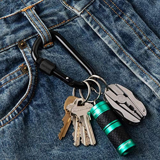 Een sleutelhanger met meerdere sleutels en een klein groen zaklampje eraan, beveiligd door een Veiligheid en veelzijdigheid onderweg: Ontdek onze hoogwaardige aluminium karabijnhaken, rustend op een blauwe denimstof.