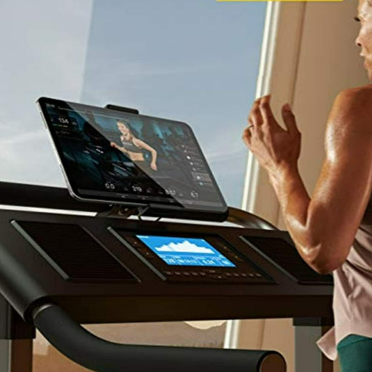 Een vrouw traint op een loopband en gebruikt een groot digitaal scherm waarop een virtuele trainingssessie wordt weergegeven, waarbij ze haar Loopband en hometrainer-tablethouder gebruikt voor handsfree entertainment.