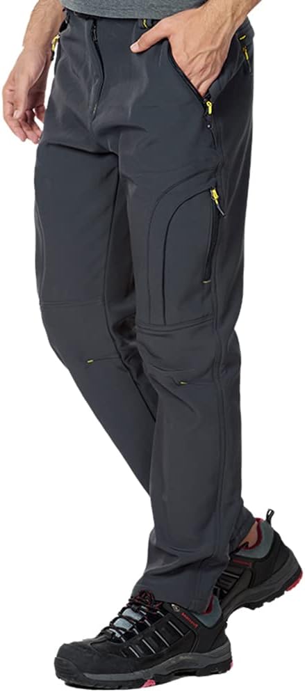 Laad afbeelding in Galerijviewer, Een man in een donkergrijze Salomon-skibroek met meerdere gele zakken met ritssluiting, gecombineerd met zwarte wandelschoenen. De broek is winddicht en ademend.
