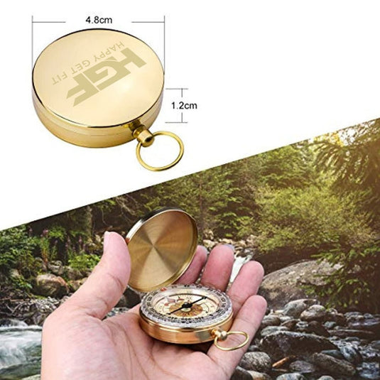 Een hand houdt een open gouden Happygetfit-zakkompas vast boven een schilderachtige rivierachtergrond, met ernaast een dimensiediagram van het gesloten kompas.