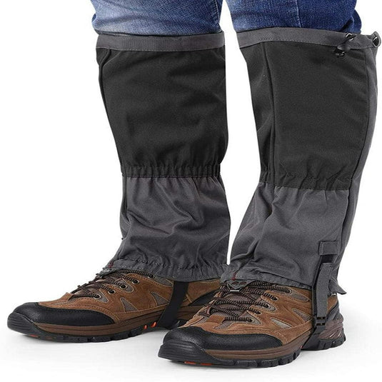 Persoon met zwarte sneeuwvangers over bruine wandelschoenen en opgerolde spijkerbroek, rechtopstaand.