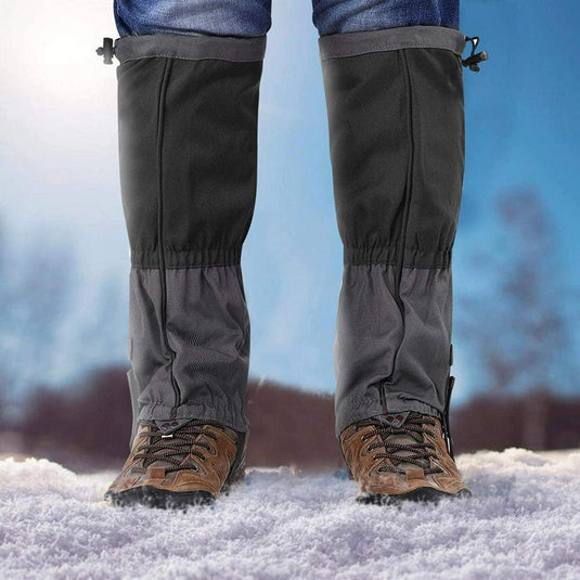 Close-up van de onderbenen van een persoon in het zwart Ontdek het ultieme comfort en bescherming met onze sneeuw gamaschen voor volwassenen en bruine wandelschoenen staande op besneeuwde grond.