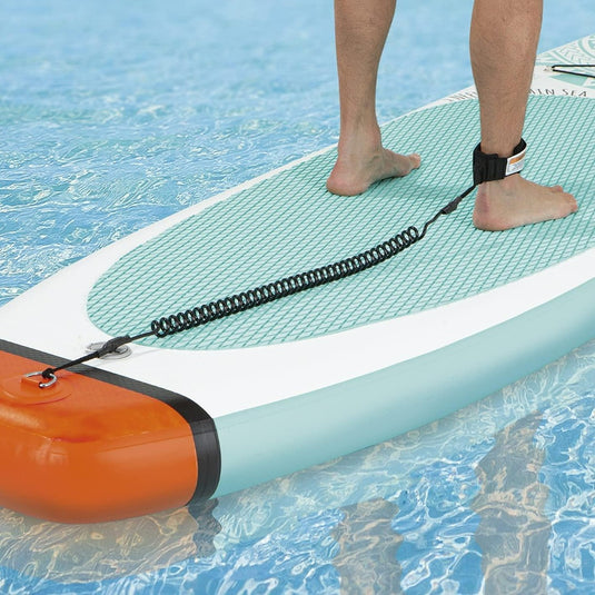 Close-up van de voeten van een persoon die op een Vitamine Sea stand-up paddleboard in een zwembad staat, met een opgerolde zwarte riem aan één enkel.