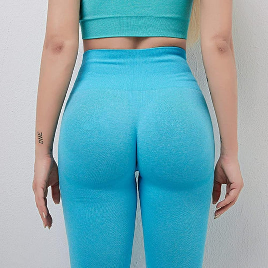 Een persoon in een turquoise crop-top en een comfortabele yoga-legging voor dames staat met zijn gezicht naar een muur, met de armen langszij, waardoor een tatoeage op de linkerarm zichtbaar is. Deze yogalegging heeft een hooggetailleerde tailleband en combineert stijl en comfort naadloos: Ontdek de perfecte balans van stijl en comfort!.