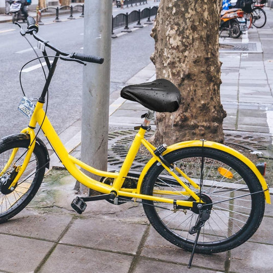 Een gele fiets met een Geniet van comfortabele fietsen met onze hoogwaardige zadelhoezen voor optimale bescherming zadelhoes vastgemaakt aan een straatpaal op een trottoir, met een nat oppervlak dat duidt op recente regen.