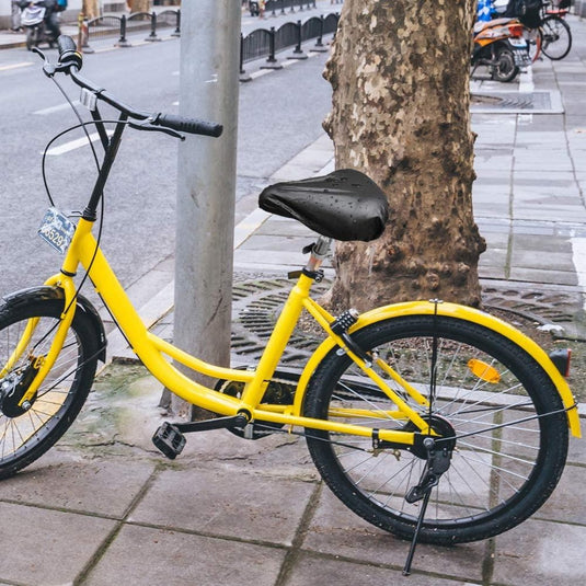 Een gele fiets met de Geniet van comfortabele fietsen met onze hoogwaardige zadelhoezen voor optimale bescherming staat geparkeerd op een nat trottoir naast een boom en paal, waarvan de fietszadel lichtjes glinstert.
