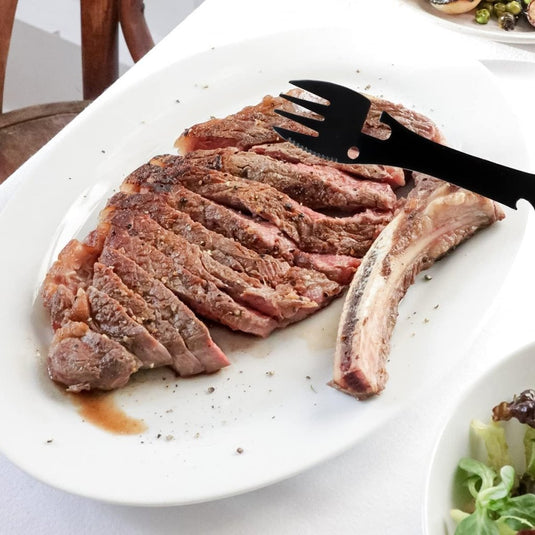 Gesneden gegrilde steak op een wit bord met een zwarte vork, perfect voor uw buitenavontuur. Naast het vlees is een bot zichtbaar, vergezeld van "De ultieme multifunctionele kampeerlepel: Jouw beste vriend voor elk avontuur.