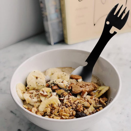 Op een marmeren oppervlak staat een kom havermout met plakjes banaan, granola en nootjes, met daarin De ultieme multifunctionele kampeerlepel: Jouw beste vriend voor elk avontuur, klaar voor een buitenavontuur. Wazige boeken zijn op de achtergrond.