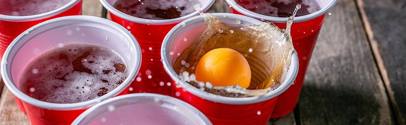 Laad afbeelding in Galerijviewer, Rode plastic bekers, waarvan één met een gele pingpongbal en opspattend vocht, staan op een houten tafel en laten het veelzijdige gebruik zien van de &quot;Speel, daag uit en knutsel met deze uitgebreide set van 100 bierpongballetjes!.
