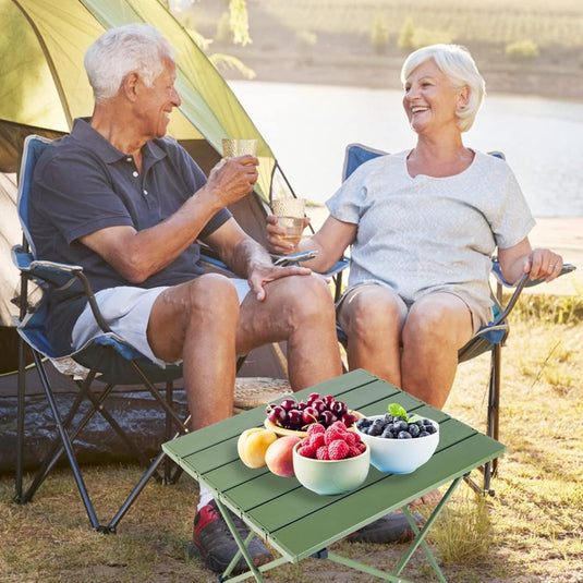 Een ouder echtpaar ontspant in kampeerstoelen, glimlachend met een drankje. Voor hen staat "Ontdek de ultieme kampeerervaring met onze draagbare campingtafel!" met een aluminium tafelblad met schalen vol appels, frambozen, kersen en bosbessen. Een tent aan het water maakt het serene tafereel compleet.