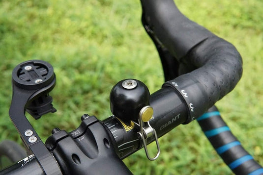 Close-up van een fietsstuur met de Stijlvol retro fietsbel en een lege apparaathouder. De getextureerde grip versterkt de klassieke uitstraling, tegen een weelderige groene grasachtergrond die de tijdloze charme versterkt.