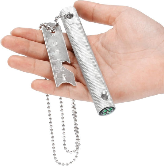 Een hand grijpt de "Robuuste vuurstarter voor outdoor avonturen," voorzien van een kompas, afneembare multitool met gekartelde rand en liniaal. Compleet met magnesium vuurstaal voor paraatheid; alle elementen sluiten naadloos aan met een kogelketting, perfect voor de outdoorliefhebber.