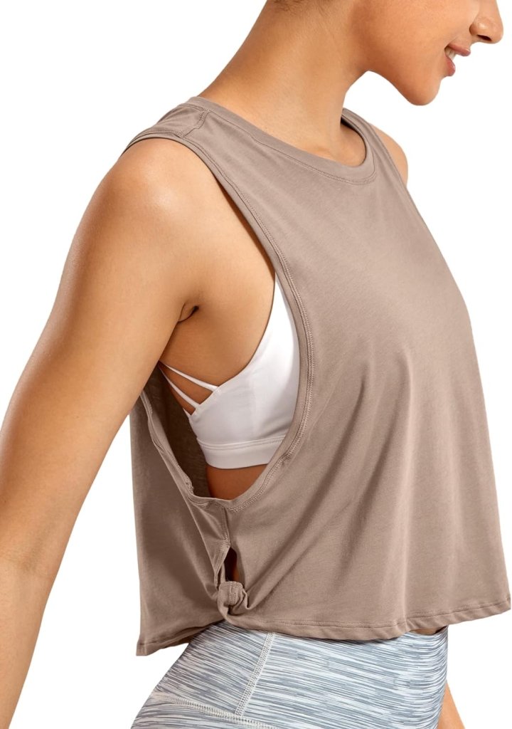 Load image into Gallery viewer, Een persoon draagt een &quot;Stijlvol en comfortabel: Yoga dames pima cotton workout tank crop&quot; in losse, mouwloze beige kleur over een witte sportbeha, gecombineerd met lichtblauwe leggings, van opzij bekeken.
