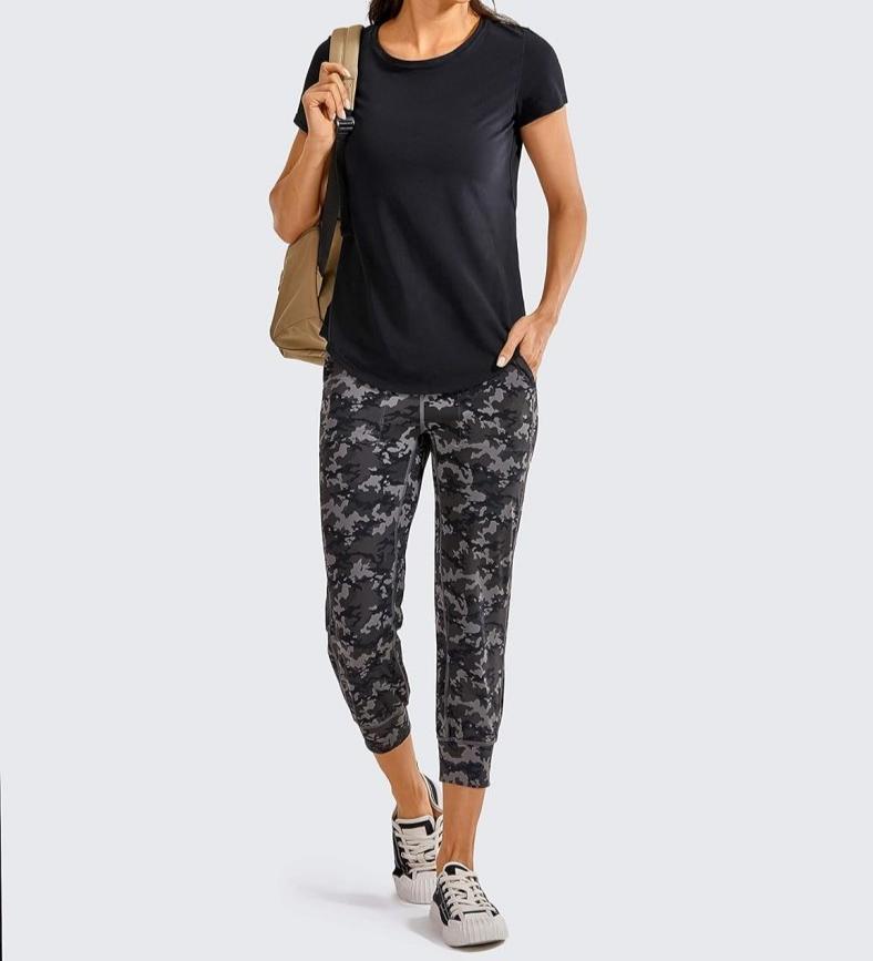 Load image into Gallery viewer, Een vrouw draagt een Perfecte yoga-T-shirt voor elke workout, een camouflagebroek en sneakers en staat zelfverzekerd met een rugzak over één schouder tegen een effen achtergrond.
