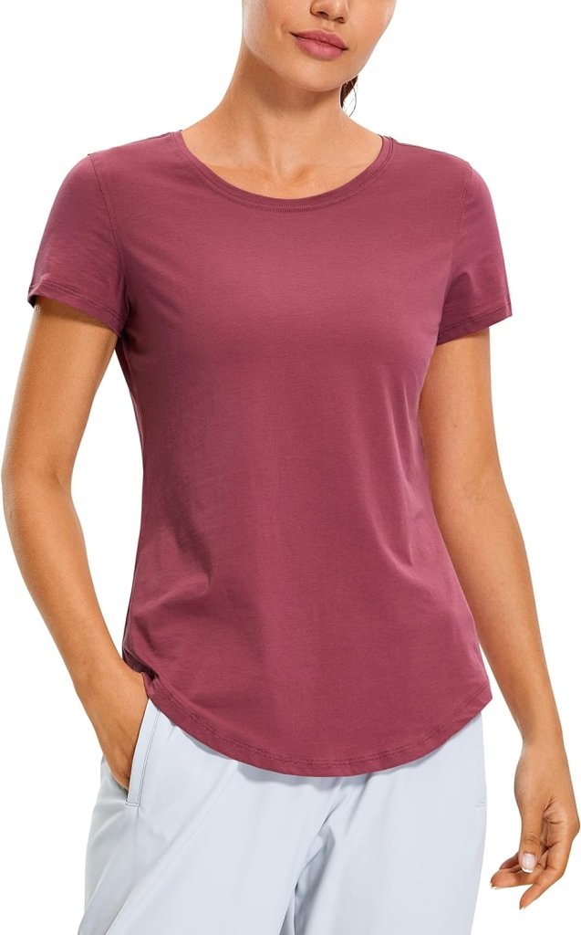 Load image into Gallery viewer, Een persoon die tijdens elke work-out het Perfecte yoga-T-shirt in de kleur mauve draagt, gecombineerd met een lichtgrijze broek, staat tegen een effen achtergrond met één hand in zijn zak.
