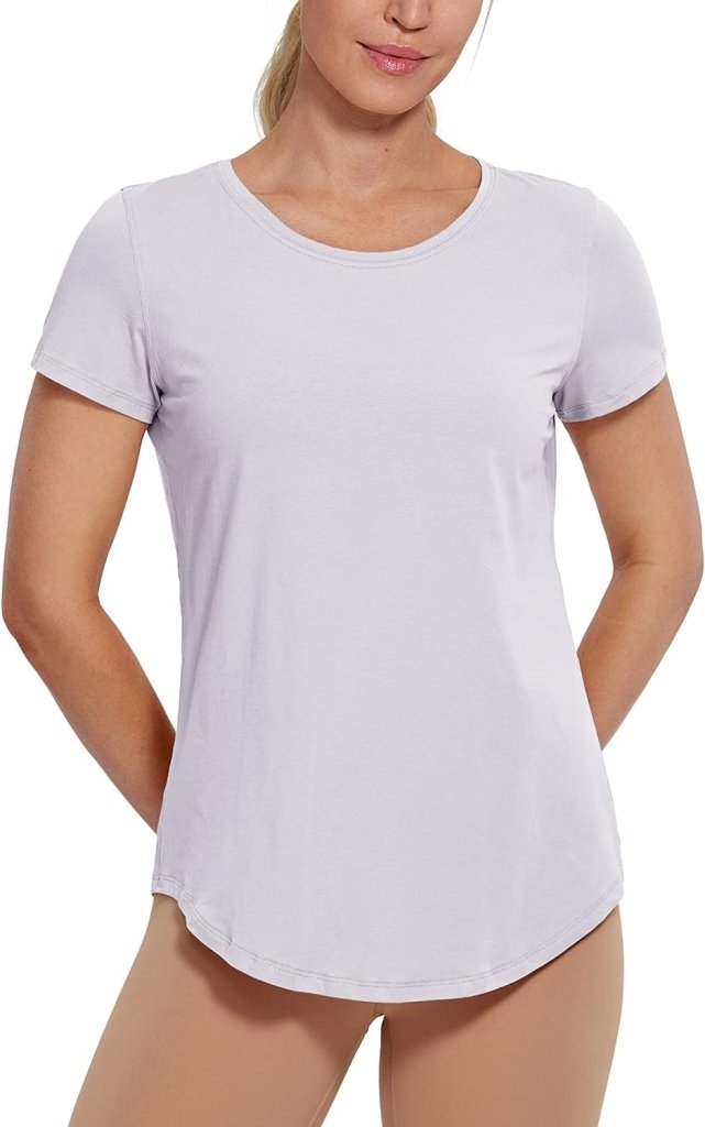 Load image into Gallery viewer, Een vrouw draagt het Perfecte yoga t-shirt voor elke workout in het wit, gecombineerd met een beige broek, en staat met haar handen op haar rug.
