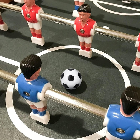 Zin met productnaam: Tafelvoetbalspel close-up met op de middenstip een mini Nooit meer zonder reserveballen tijdens een tafelvoetbalwedstrijd, met rode en blauwe spelersfiguren klaar om te spelen.